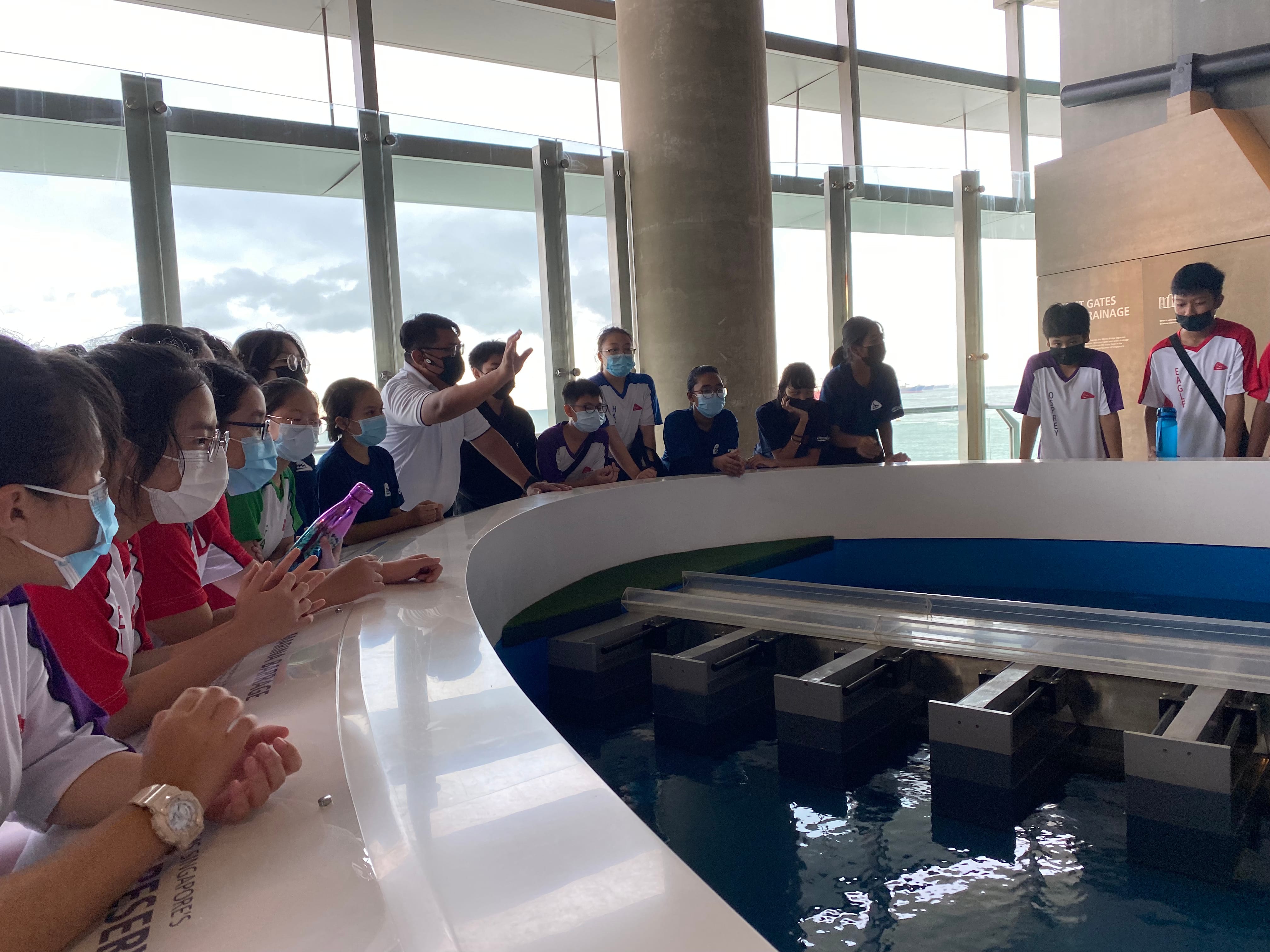 Students wearing masks gather around a circular water exhibit indoors while a guide points and explains.